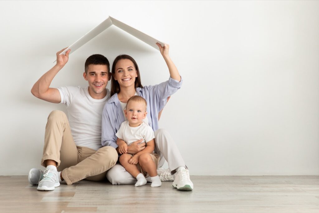 Family Housing. Happy Parents With Cute Infant Baby Sitting Under Cardboard Roof
