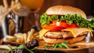 American cuisine concept. Juicy burger with meat patty, tomatoes, cheddar cheese, lettuce and homemade bun. In the background are french fries and a glass of beer. Close up